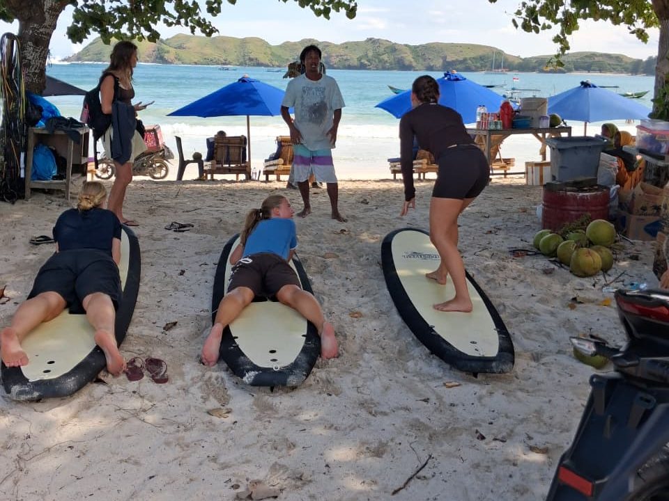 A beach lesson: three students practising pop-ups on soft-tops while the coach demonstrates, the bay behind them